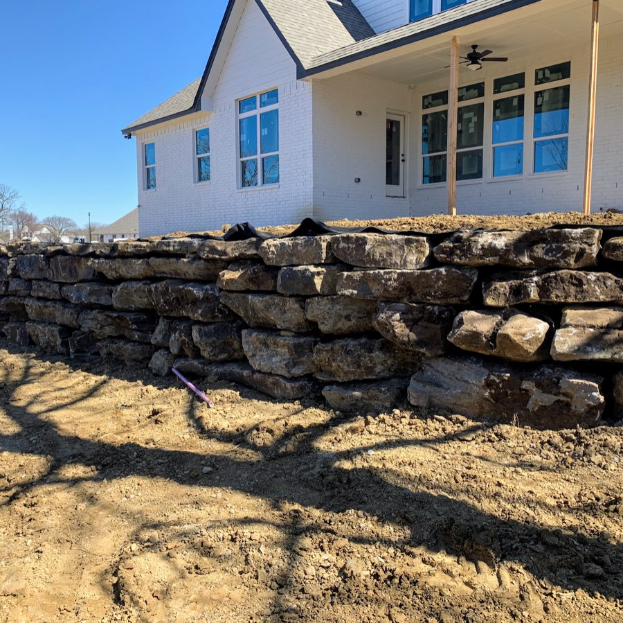 Long stacked-stone retaining wall in front of a white brick home, freshly built